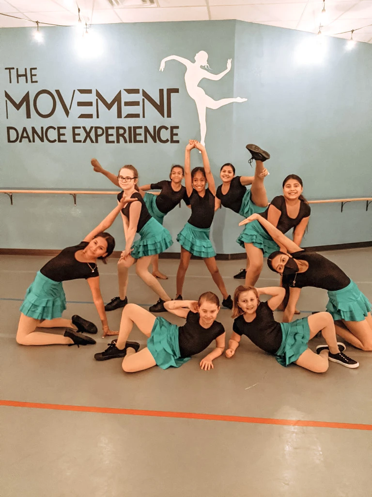 Movement Dance Company dancers in black tops and green skirts striking a group pose in the studio